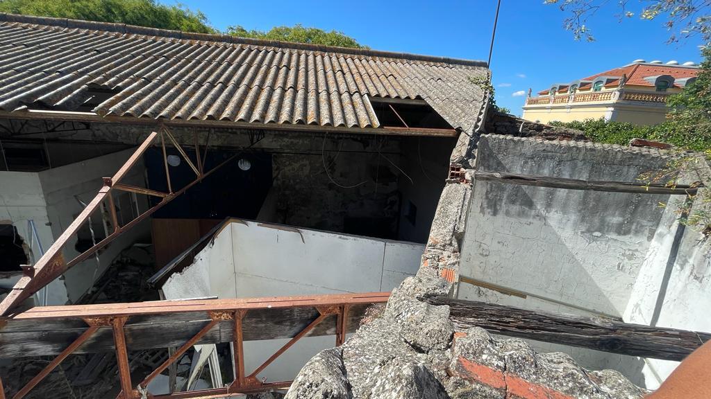 Telhado de fibrocimento de um armazém abandonado, visto do alto: parte da cobertura já ruiu sobre o interior, deixando à vista asnas metálicas enferrujadas, paredes caiadas e entulho, com um edifício amarelo de telhado vermelho ao fundo.