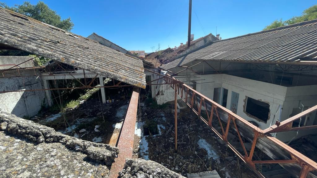 Vista do alto sobre o pátio interior de um conjunto de armazéns devolutos: asnas metálicas enferrujadas atravessadas sobre o vazio, coberturas partidas, paredes brancas com vãos rasgados e entulho no chão.