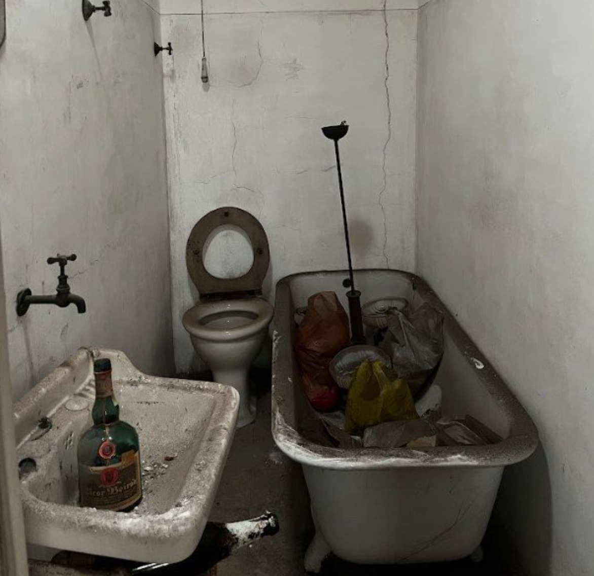 Casa de banho devoluta e degradada: lavatório manchado com uma garrafa de licor antiga, sanita com tampo de madeira e banheira de ferro cheia de sacos de lixo.