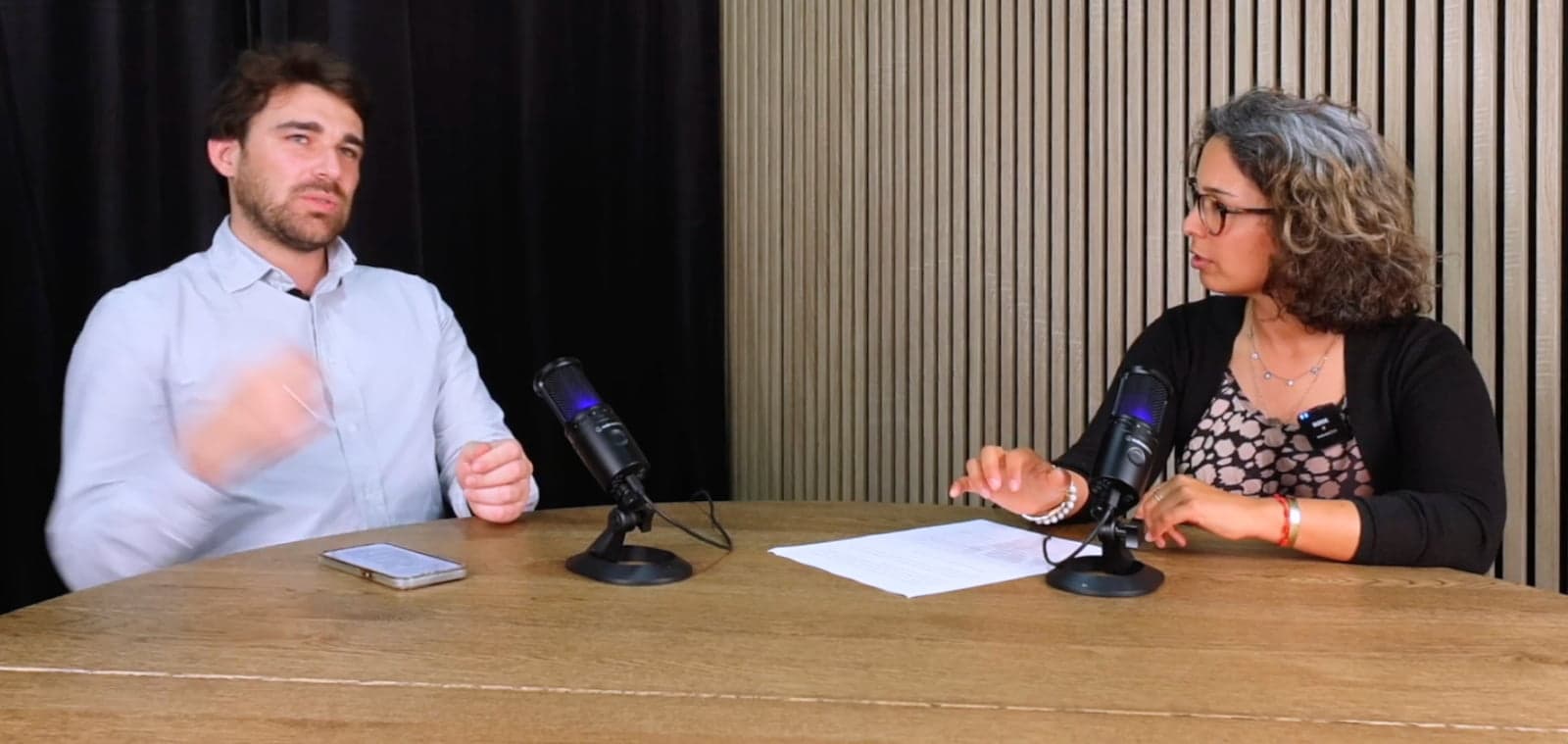 Um homem e uma mulher sentados a uma mesa de madeira com dois microfones de podcast, papéis e um telemóvel, diante de uma parede de ripas de madeira.