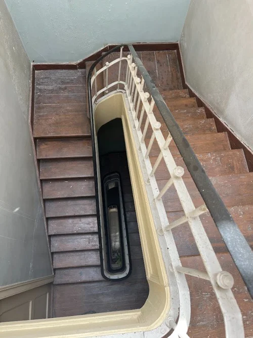 Caixa de escadas de um prédio antigo vista de cima, com degraus de madeira escura muito gastos, corrimão e guarda de ferro pintados de creme e a bomba de um elevador instalado no vazio da escada.