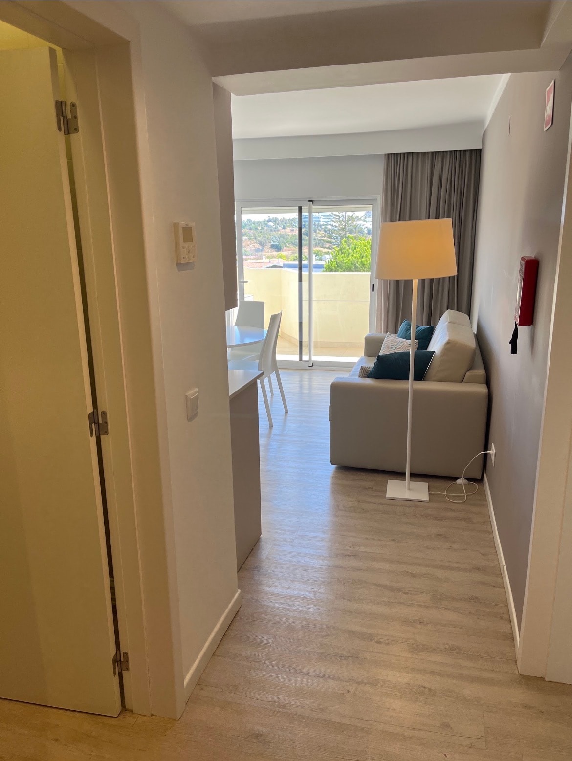 Corredor de entrada de um apartamento turístico a dar para a sala: sofá bege com almofadas azuis, candeeiro de pé aceso, mesa de refeições branca e porta de correr envidraçada para a varanda; na parede, um extintor e um comando de ar condicionado.