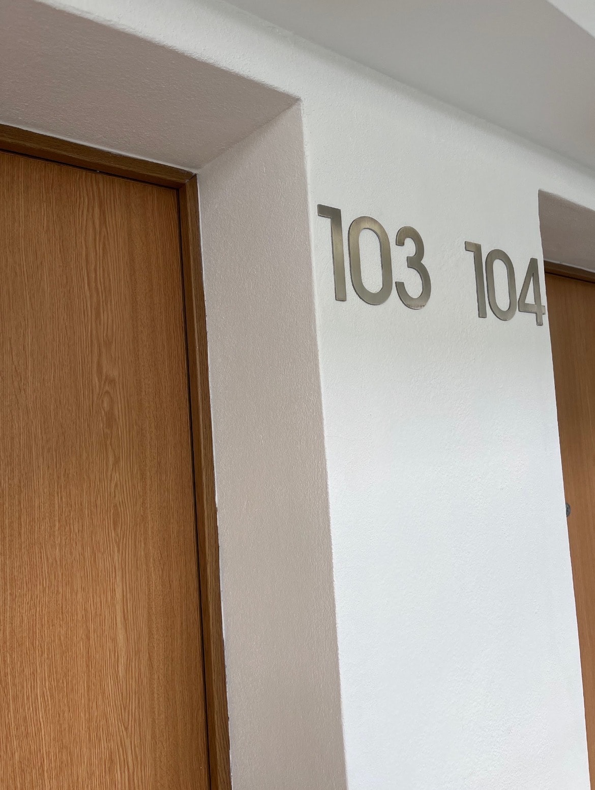 Corredor de um edifício com paredes brancas e portas em madeira clara, com os números de quarto 103 e 104 em metal escovado aplicados na parede.