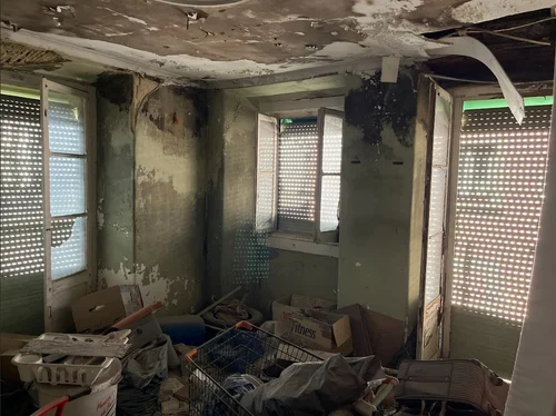 Divisão devoluta e cheia de tralha, com o tecto queimado e a descascar, paredes com o reboco a cair, e três janelas com portadas de réguas por onde a luz entra em riscas; no chão, caixotes, sacos e uma grade de metal.