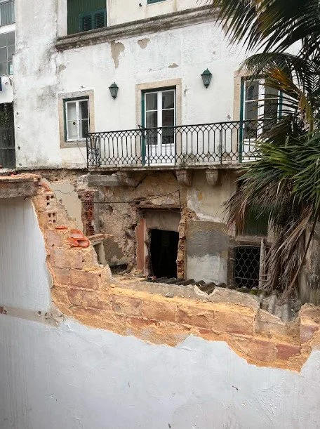 Empena de um prédio antigo com o reboco arrancado a descobrir a alvenaria de pedra e tijolo e um vão tapado, tendo por cima uma varanda de ferro forjado e as janelas de guilhotina do edifício vizinho, com uma palmeira à direita.