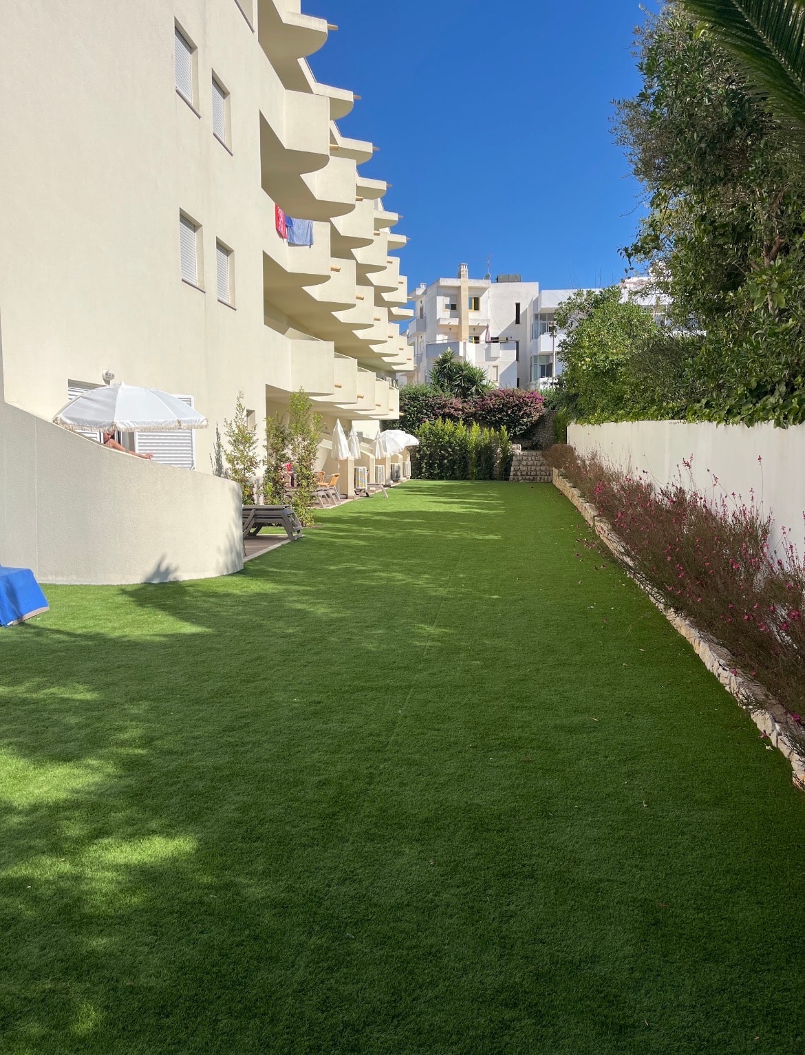 Jardim de relva sintética entre um aparthotel de varandas em consola e um muro caiado com canteiro florido, com chapéus de sol e espreguiçadeiras junto ao edifício e o casario branco da vila ao fundo.