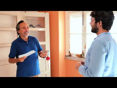 Dois homens de pé numa divisão em obras, de paredes cor de laranja e caixilharia antiga; o mais velho segura folhas de papel enquanto fala.