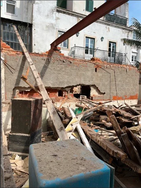 Obra de demolição no logradouro de um prédio: uma viga metálica vermelha escorada sobre um muro de alvenaria rasgado, escadas de madeira encostadas e um monte de vigamento e tábuas arrancadas em primeiro plano.