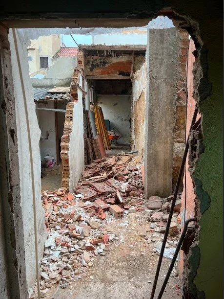 Vista através de um vão aberto à picareta para o interior demolido de um edifício: corredor estreito coberto de tijolos e telhas partidas, paredes descarnadas até ao tosco e tábuas de madeira encostadas ao fundo.