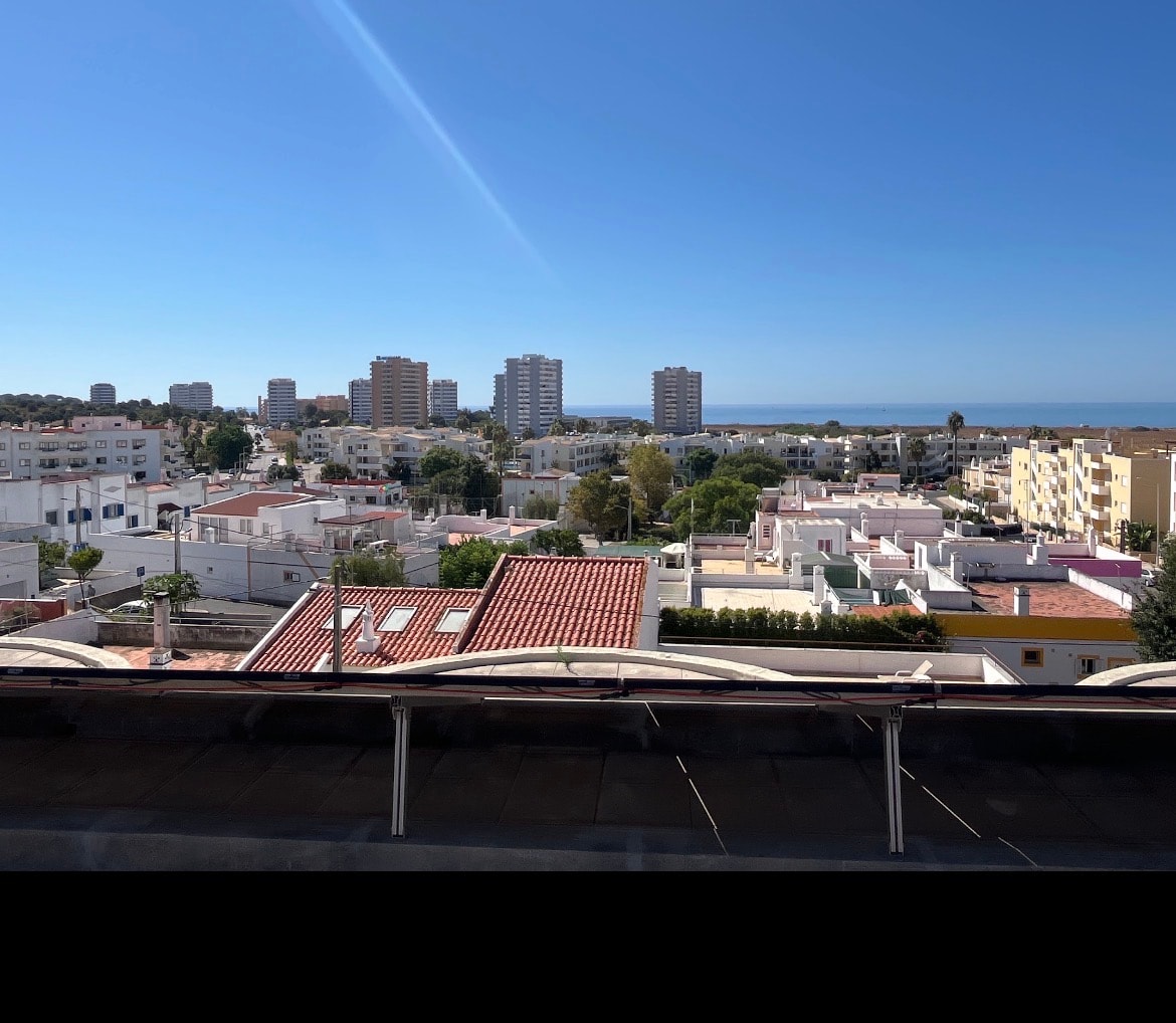 Vista de uma cobertura sobre uma povoação do litoral algarvio: telhados de telha e terraços brancos em primeiro plano, torres de apartamentos ao fundo e a linha do mar no horizonte, sob céu limpo.